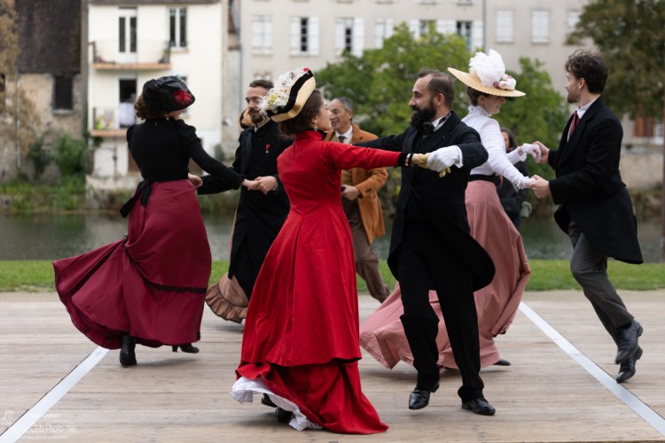 Danse reconstitution historique : Un groupe de personnes en tenues d'époque 1900 dansent en rond sur un parquet au bord d'une rivière.